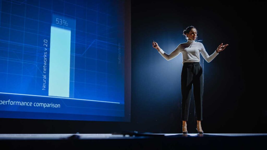 woman talking on a stage infront of a projection of a bar graph