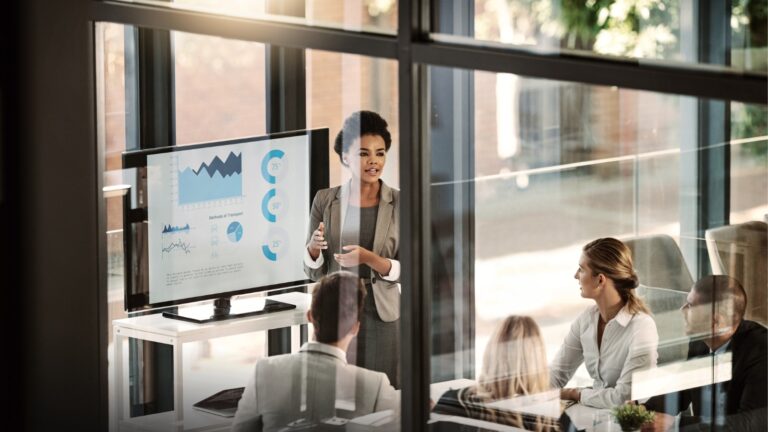 woman of color presenting to room of people in windowed office