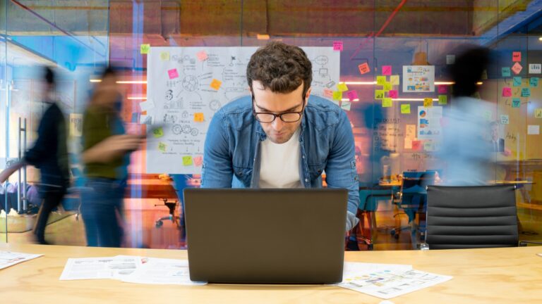 Man working at a creative office using his computer and people moving at the background